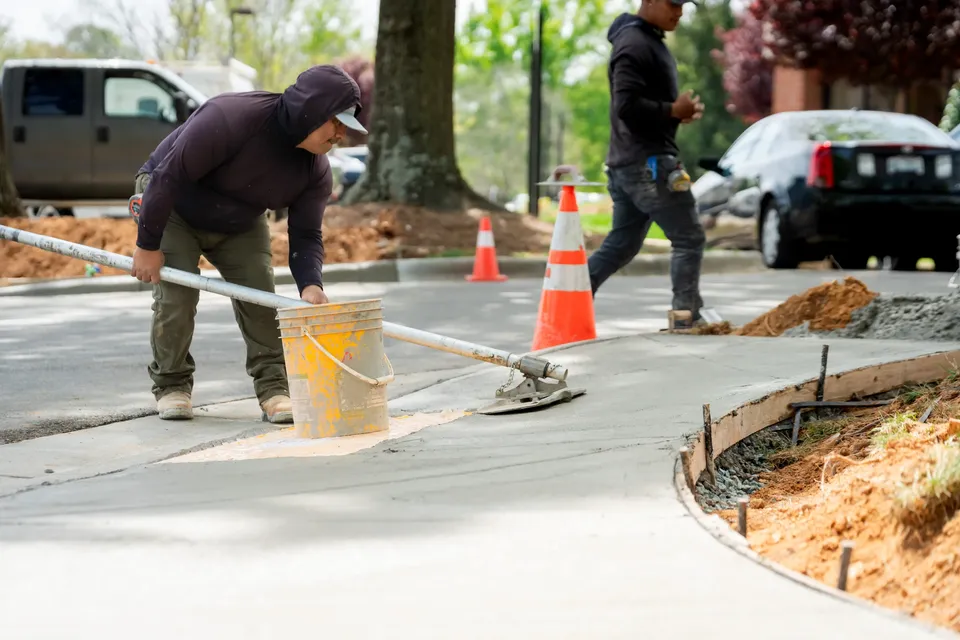Crew member smoothing a freshly poured concrete sidewalk beside traffic cones.
